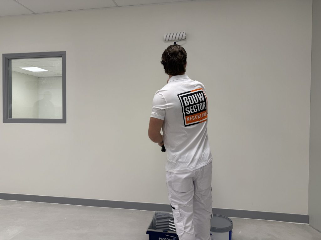 Schilder van Bouwsector Nederland rolt een witte muur met verfbak en emmer op de betonvloer