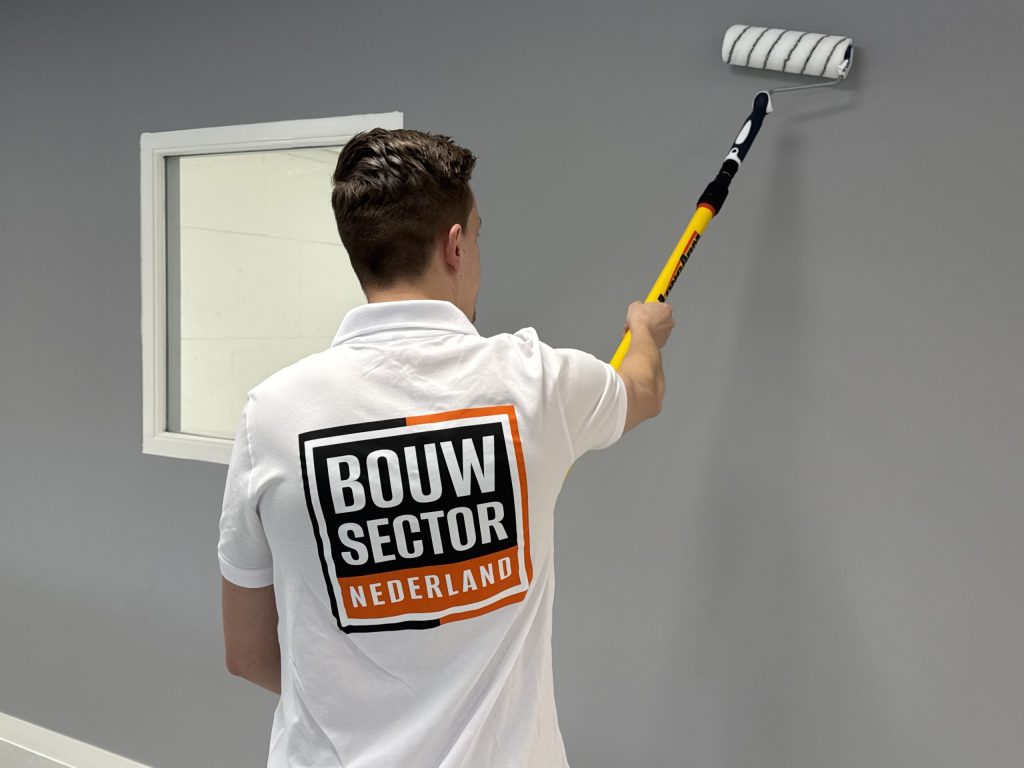 Schilder met het Bouwsector-logo op zijn rug rolt een grijze muur naast een binnenraam met witte lijst
