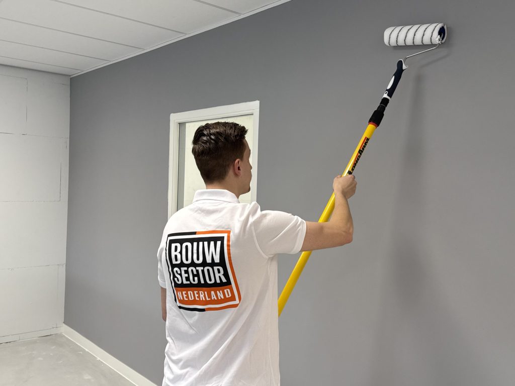 Schilder van Bouwsector Nederland zet een muur in een grijze kleur met een roller aan een gele steel