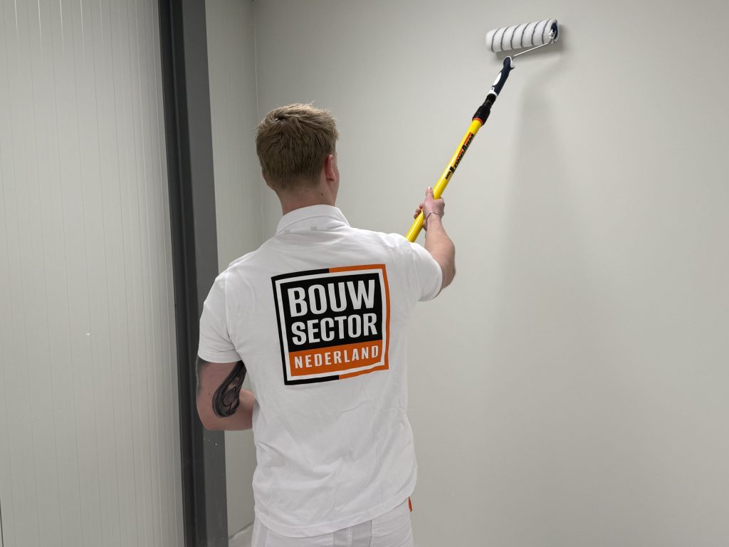 Schilder in wit shirt met Bouwsector-logo zet een lichte wand in de verf naast een deurpost
