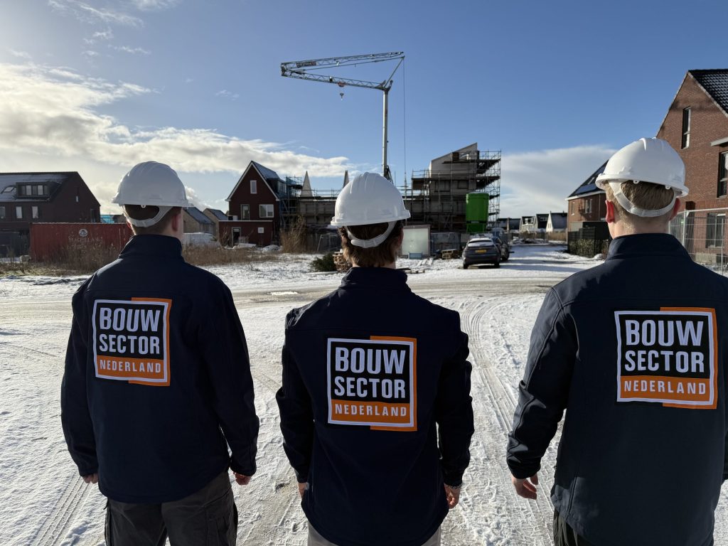 Drie vakmensen van Bouwsector Nederland met witte helm en donkere werkjas, van achteren gezien, voor woningen in aanbouw