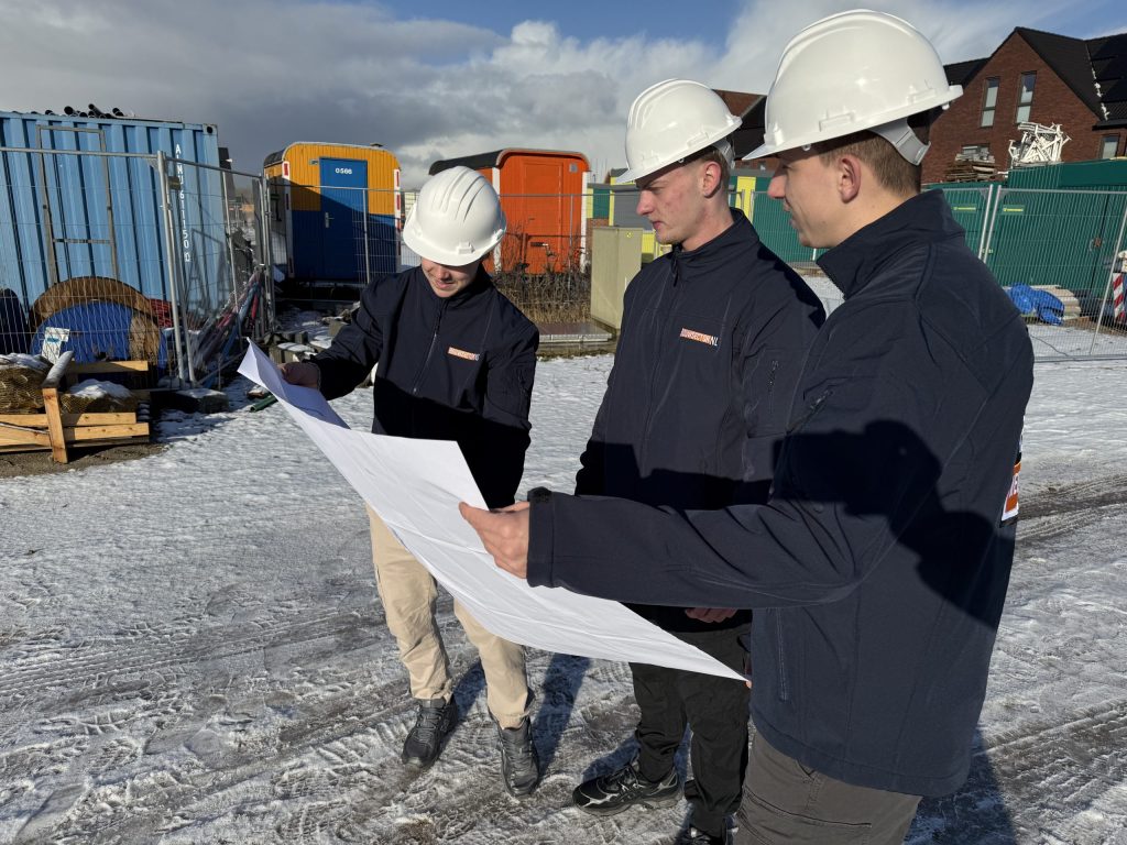 Drie vakmensen van Bouwsector Nederland met witte helm bekijken samen een bouwtekening buiten in de sneeuw