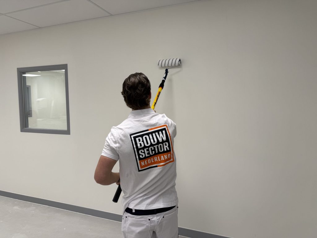 Schilder rolt een witte wand onder een plafond van systeemplaten, naast een binnenraam met grijs kozijn